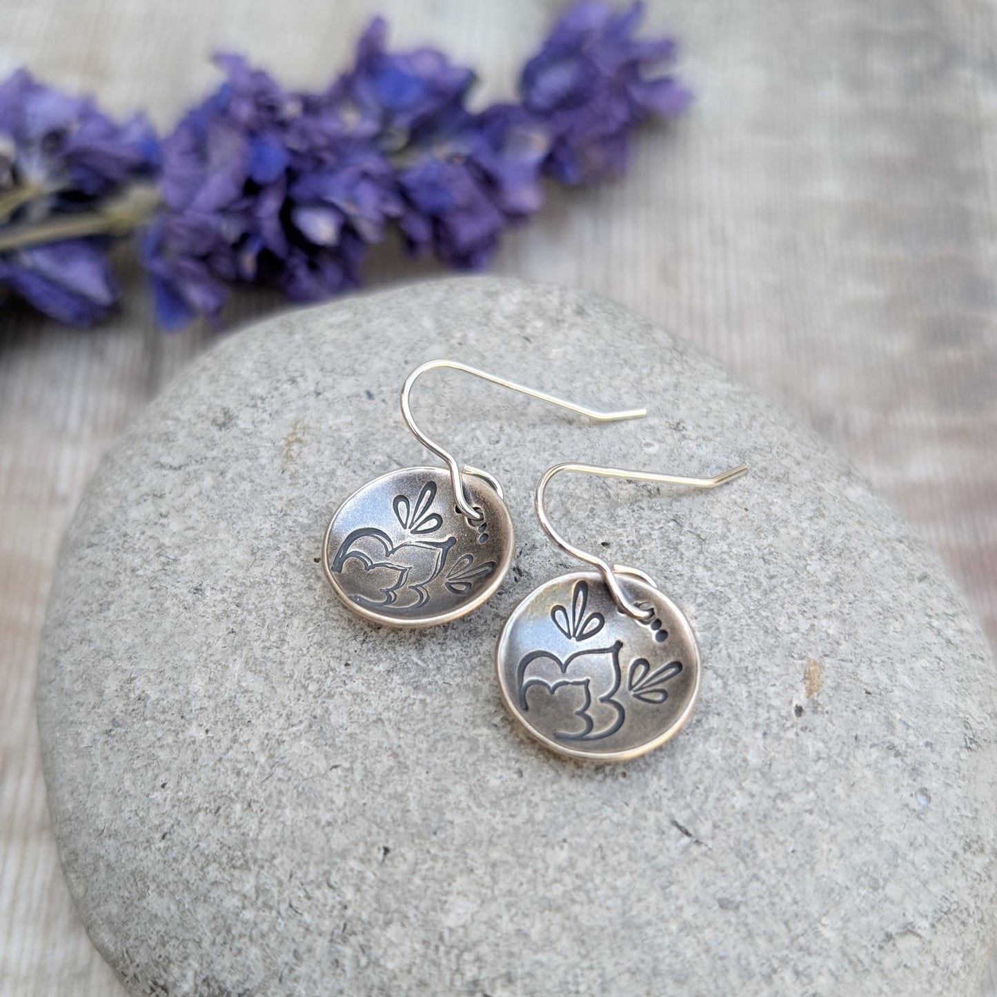 Silver floral earrings on a stone with purple flowers in the background
