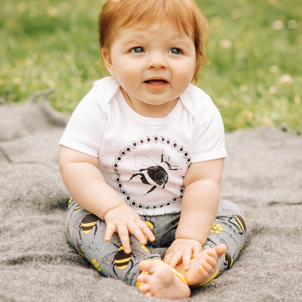A baby sitting outdoors on a gray blanket wearing a white short-sleeve bodysuit with a black bee graphic and gray patterned pants with yellow accents.