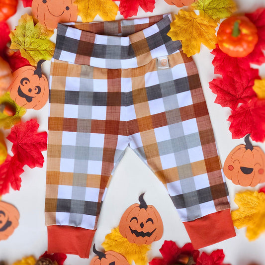 A pair of toddler plaid leggings with orange cuff accents, laid flat on a white background surrounded by autumn-themed decorations including paper pumpkins, red and yellow leaves, and small faux pumpkins.