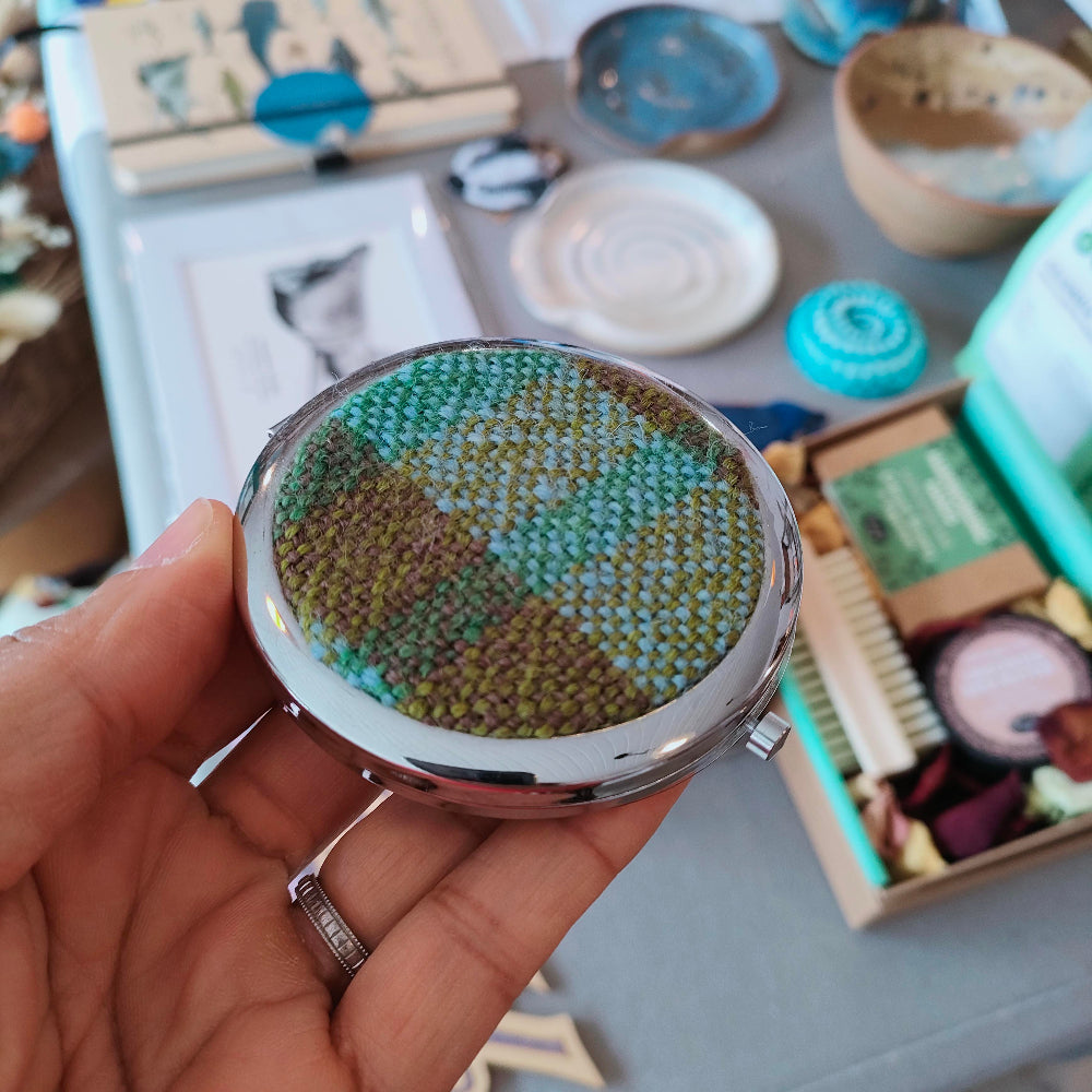 Hand holding a compact mirror with a textured green and brown design, surrounded by various items on a table.