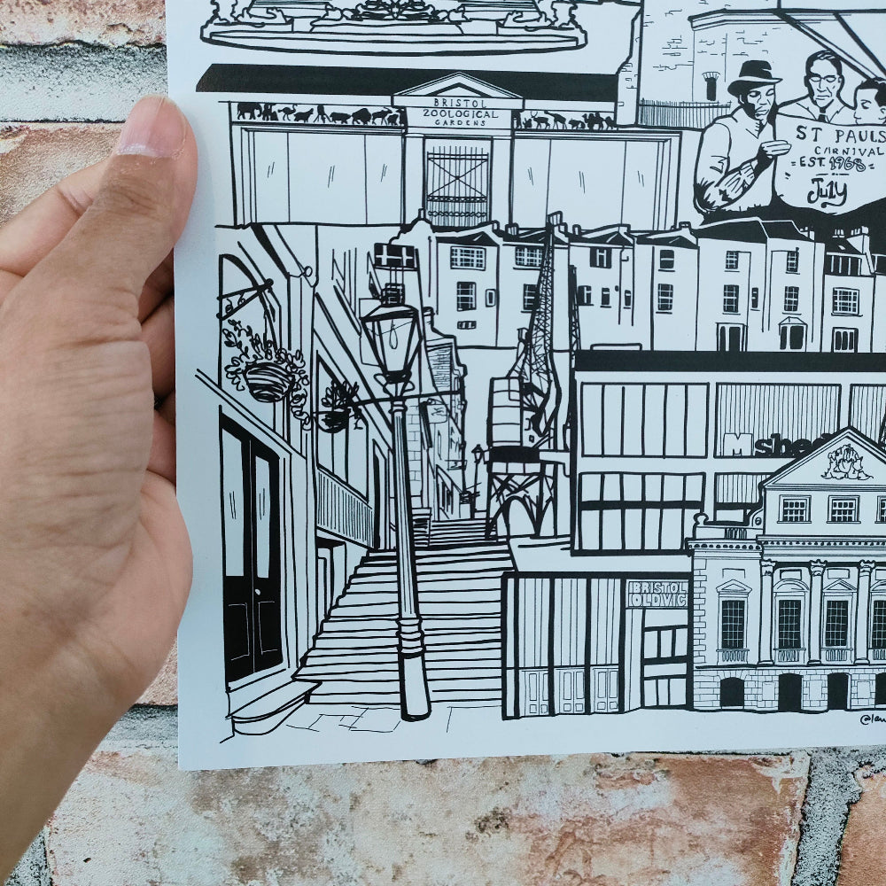 A hand holds a printed black-and-white street scene illustration featuring buildings, a staircase, a lamppost, and a small inset of two men with text reading 'ST. IAN'S DAY 1978'.
