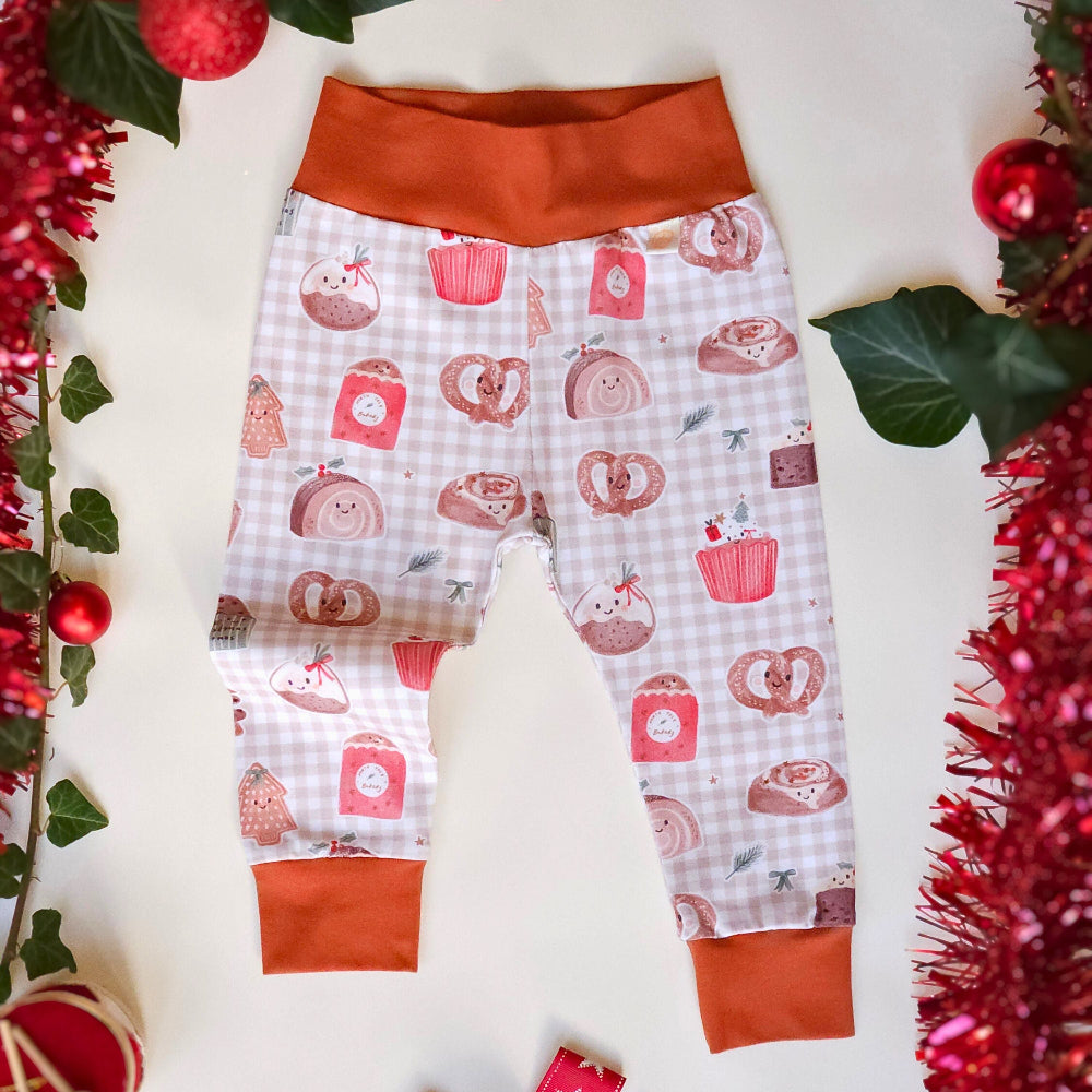 A pair of baby leggings with a white gingham pattern featuring red and pink holiday treats like cupcakes, pretzels, and cinnamon rolls, finished with solid rust-orange waistband and cuffs. The leggings are displayed in a flat-lay setting surrounded by festive Christmas decorations including red tinsel, ornaments, and a red star-patterned bow.