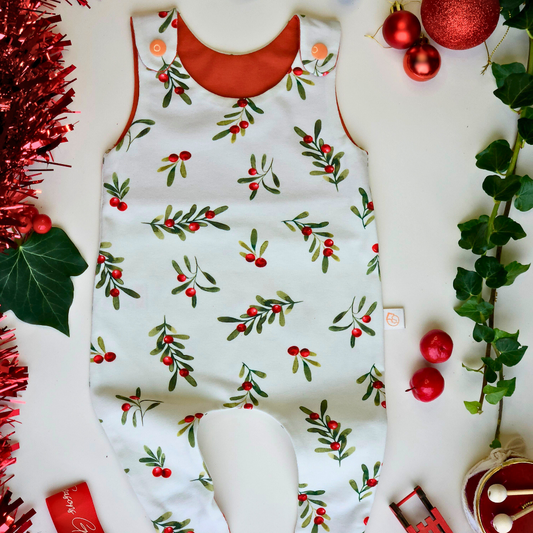 A baby jumpsuit laid flat on a white surface, decorated with a green mistletoe and red berry pattern. The jumpsuit has orange trim along the neckline and leg openings, and orange buttons at the shoulder straps.