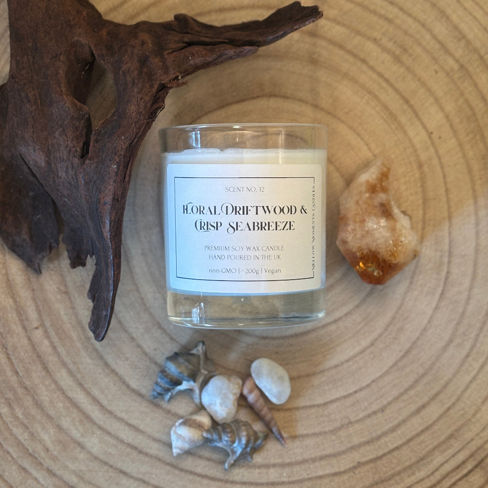 A glass jar candle labeled 'Floral Driftwood & Crisp Seabreeze' by Scents of the Coast, styled on a wood slice with driftwood, shells, and a small crystal.