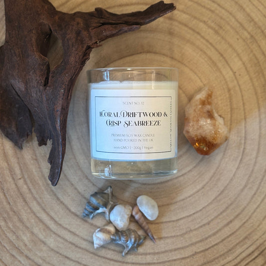 A glass jar candle labeled 'Floral Driftwood & Crisp Seabreeze' by Scents of the Coast, styled on a wood slice with driftwood, shells, and a small crystal.