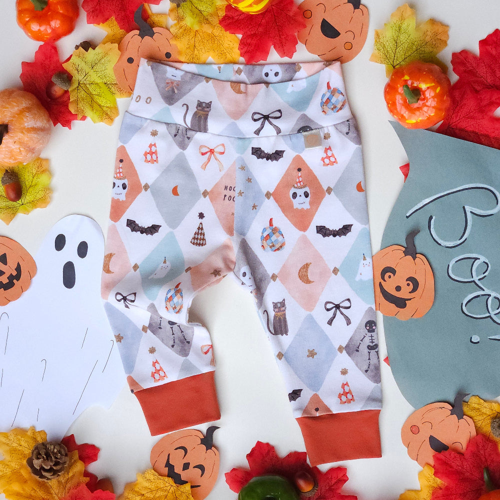A pair of baby leggings with a Halloween-themed print featuring bats, cats, pumpkins, and bows on a light gray and peach diamond pattern, styled in a flat lay with autumn leaves, pumpkins, and a ghost cutout.