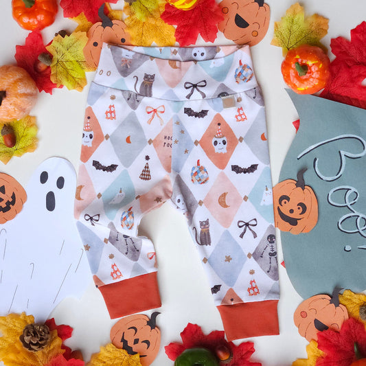 A pair of baby leggings with a Halloween-themed print featuring bats, cats, pumpkins, and bows on a light gray and peach diamond pattern, styled in a flat lay with autumn leaves, pumpkins, and a ghost cutout.