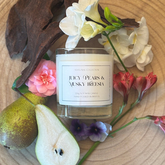 Candle labeled 'Juicy Pears & Musky Freesia' surrounded by pears and flowers on a wooden surface