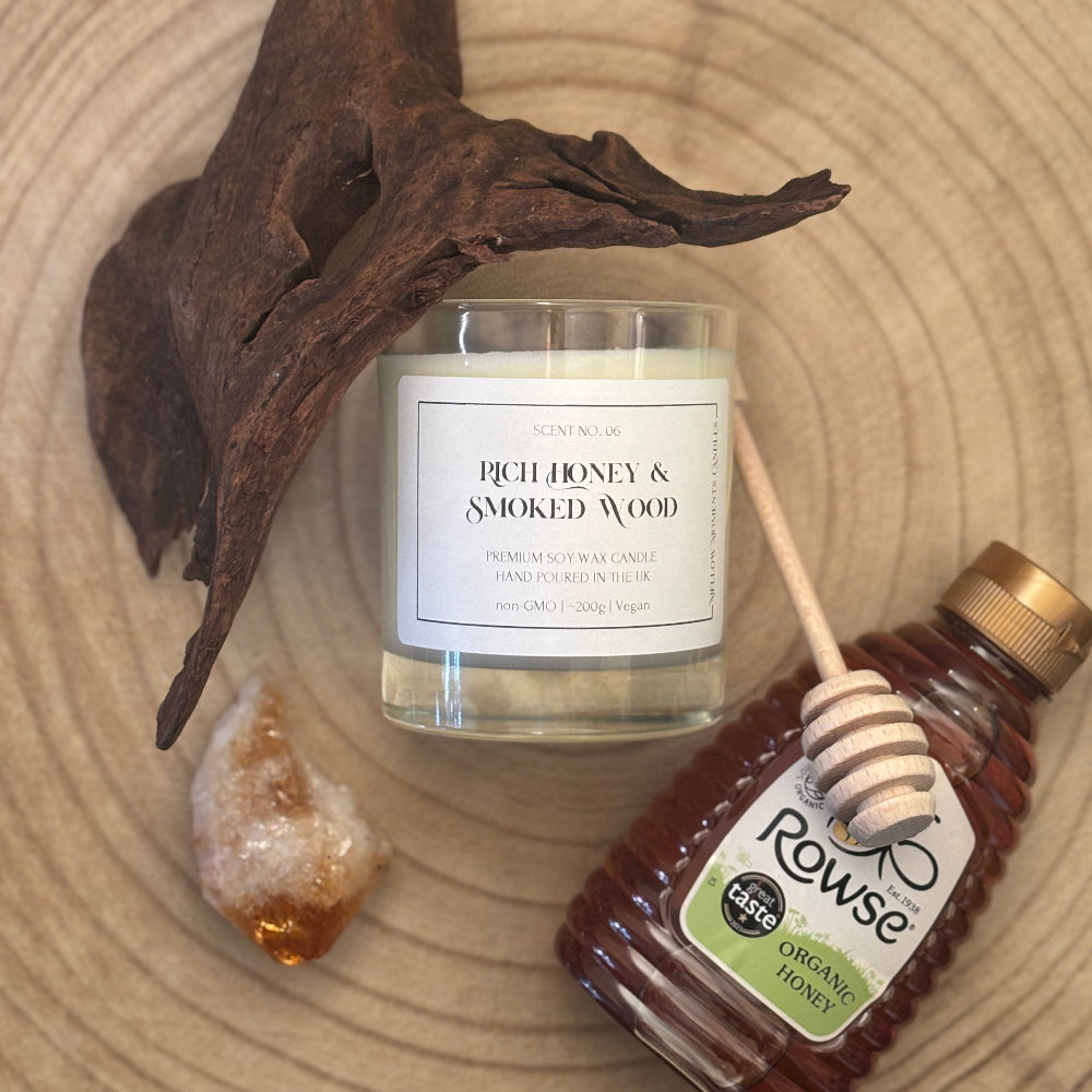 A candle jar labeled 'RICH HONEY & SMOKED WOOD' is styled on a wood slice with a dried branch, a honey dipper, a small crystal, and a bottle of Rowse Organic Honey.