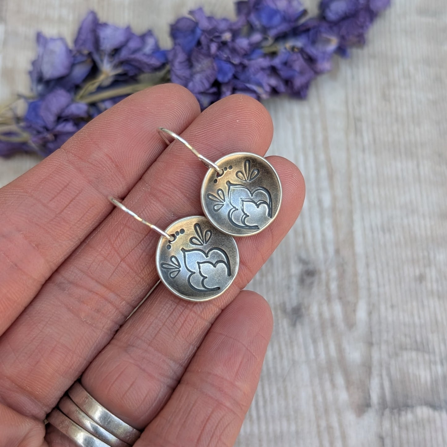 Silver earrings with floral designs held between fingers against a textured background with purple flowers.