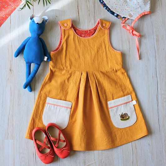 A baby girl's mustard yellow linen pinafore dress with white front patch pockets, laid flat on a light wood floor alongside a blue plush toy, a white polka-dot sun hat, and red ballet flats.