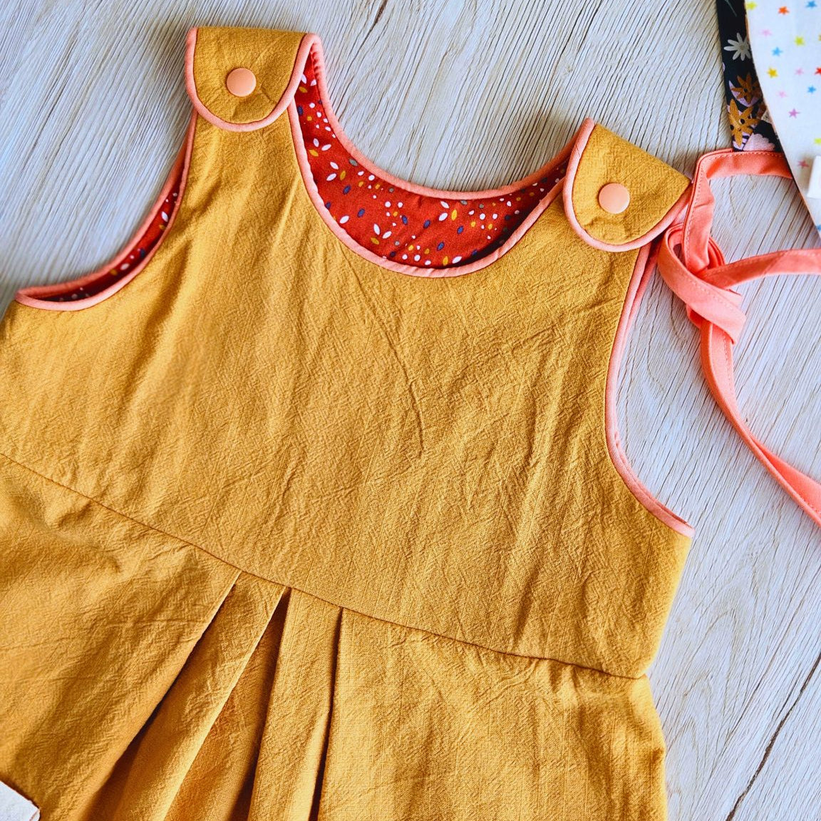 A mustard yellow sleeveless baby dress with a red patterned inner collar and pinkish-red trim, laid flat on a light wood background.