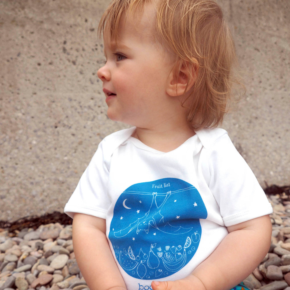 A baby wearing a white short-sleeve bodysuit with a blue graphic of a baby carriage, stars, and pears, sitting outdoors on a pebble ground against a concrete wall.