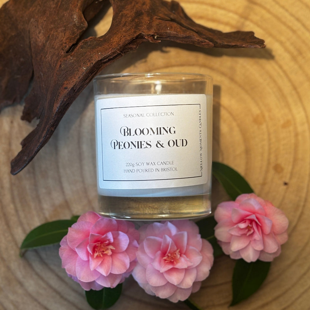 Candle labeled 'Blooming Peonies & Oud' surrounded by pink flowers on a wooden surface