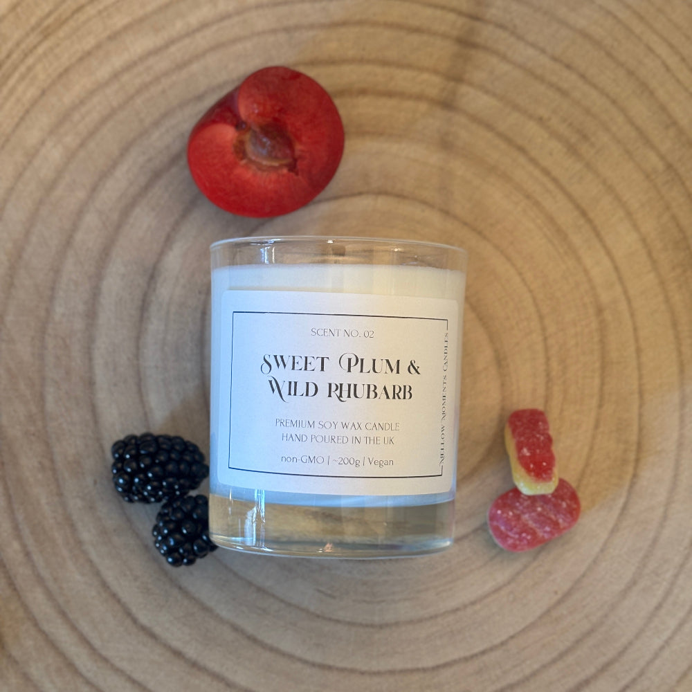 A glass jar candle labeled 'Sweet Plum & Wild Rhubarb' sits on a round wooden surface, surrounded by a halved plum, blackberries, and pieces of rhubarb.